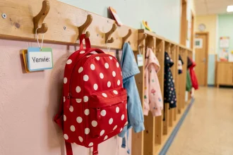 Petit sac à dos maternelle rouge à pois blancs accroché sur un porte-manteau en bois dans un couloir d'école